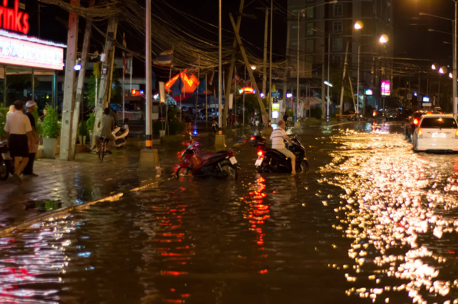 A flooded street in Pattaya at night after heavy rain, with motorbikes riding through standing water under the street lights