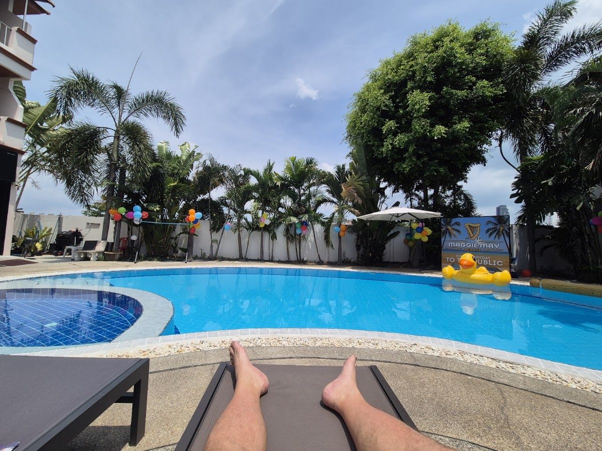 Maggie Mays Resort Jomtien Pattaya — the outdoor pool surrounded by palm trees, sun loungers and a Maggie May resort sign visible poolside